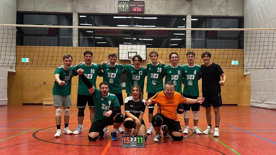 Volleyballmannschaft posiert nach dem Spiel, alle in gr&uuml;nen Trikots, mit Ergebnistafel 15:11 auf dem Boden.