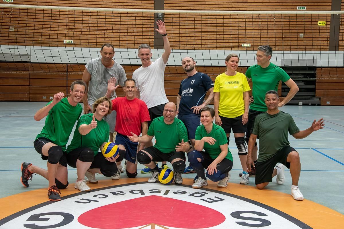 Gruppe von Volleyballspielern in bunter Sportkleidung posiert l&auml;chelnd auf dem Spielfeld vor einem Netz.