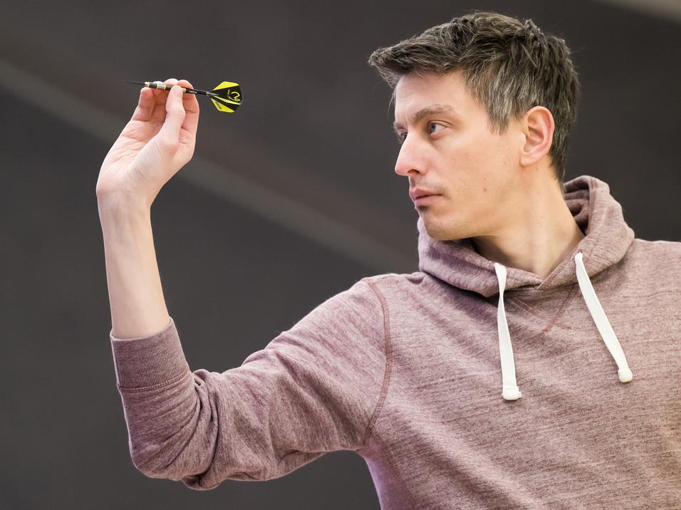 Ein Mann in einem grauen Hoodie zielt mit einem Dart auf eine Dartscheibe in einer Turnierumgebung.