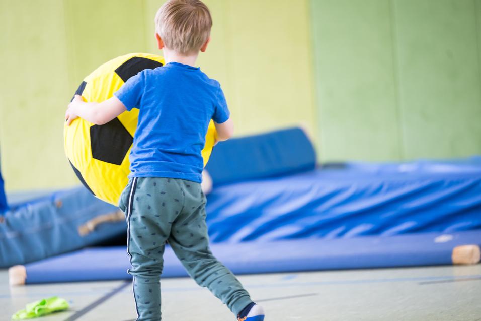 Ein Junge trägt einen großen, gelben Ball mit schwarzen Feldern in einer Turnhalle.