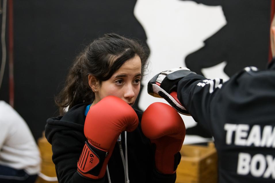 Junge Boxerin in roter Ausrüstung konzentriert auf Training mit Trainer im Hintergrund, Boxhandschuhe bereit.