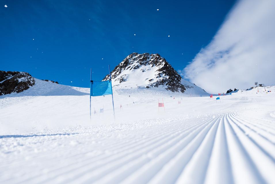 Schneebedeckte Piste mit blauen und roten Toren, im Hintergrund ein schneebedeckter Berg unter blauem Himmel.
