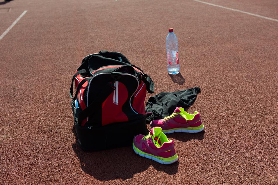 Sporttasche, pinke Sportschuhe, schwarze Kleidung und eine Wasserflasche auf einer orangefarbenen Laufbahn.
