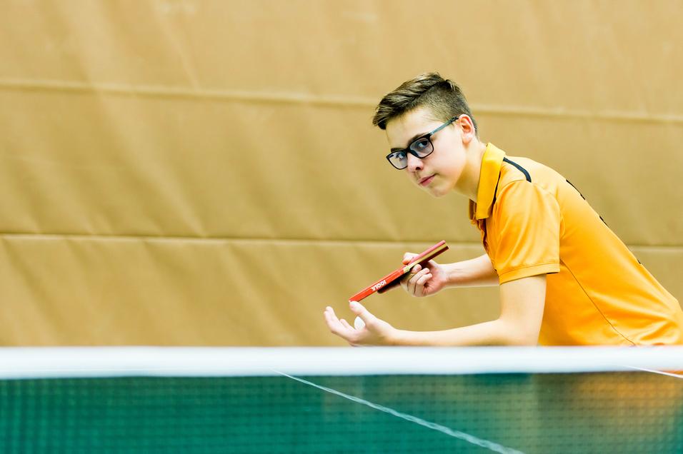 Ein junger Tischtennisspieler in orangem Shirt bereitet sich mit Schläger in der Hand auf den Aufschlag vor.