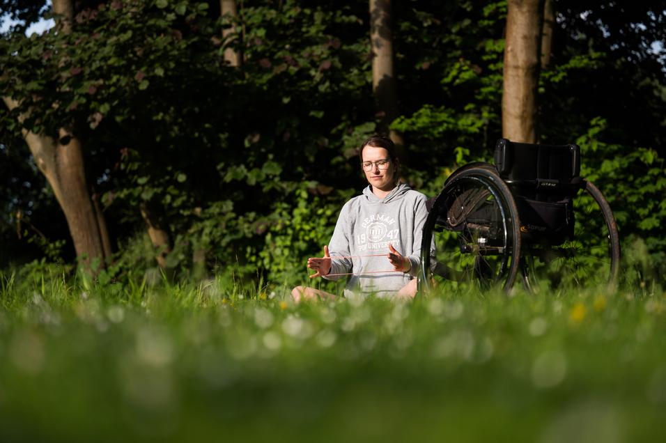 Junge Frau in einem grauen Hoodie sitzt im Gras und meditiert, ein Rollstuhl steht neben ihr.